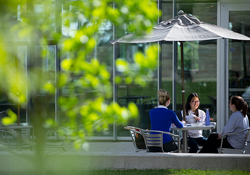 Students sitting and talking under a table with a patio umbrella on Brescia's grounds..